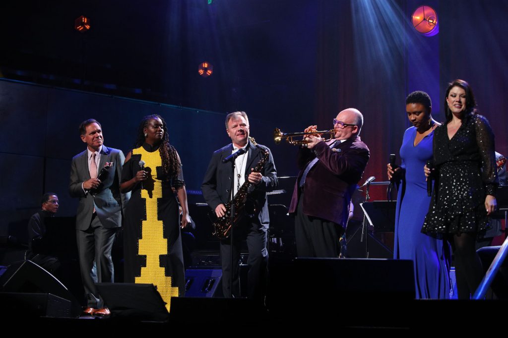 A man plays the piano while six people stand on a dark stage singing and playing musical instruments